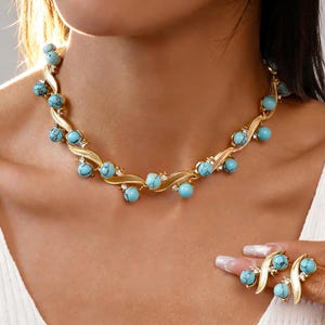 Pode incluir: Um colar dourado e um conjunto de brincos a condizer. O colar e os brincos apresentam contas de cor turquesa e pequenos cristais transparentes. A joalharia é desenhada com um padrão fluido em forma de folha.