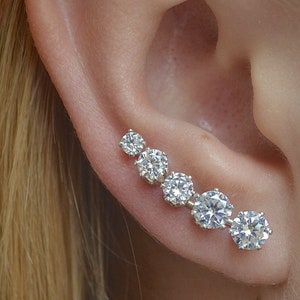 May include: A silver ear cuff with five clear cubic zirconia stones set in a graduated size, starting with the smallest stone closest to the earlobe and increasing in size as the stones move up the ear.