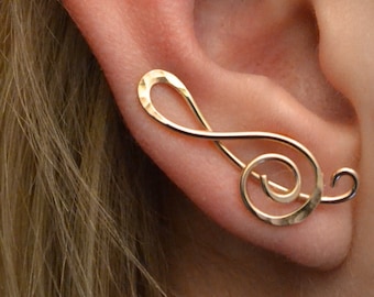 May include: Gold ear cuff in the shape of a treble clef. The cuff is worn on the upper part of the ear.