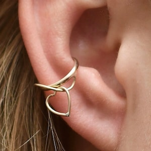 May include: Gold-colored ear cuff featuring a heart-shaped design. The cuff wraps around the ear and has a delicate, open heart pendant. The jewelry is worn on a person's ear, with the skin tone visible.