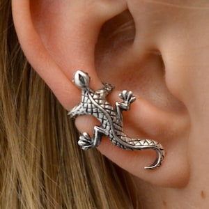May include: A silver-toned lizard ear cuff. The cuff wraps around the ear, with the lizard's body and tail curving along the ear's edge. The lizard has detailed scales and claws, creating a realistic appearance.
