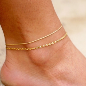 Pode incluir: Duas tornozeleiras douradas no tornozelo de uma pessoa. As tornozeleiras são sobrepostas, com uma corrente fina sobre uma corrente de corda mais grossa.
