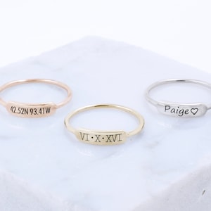 May include: Three delicate gold rings with engraved inscriptions. The rings are on a white surface. The inscriptions are "42.52N 93.41W", "VI.X.XVI", and "Paige".