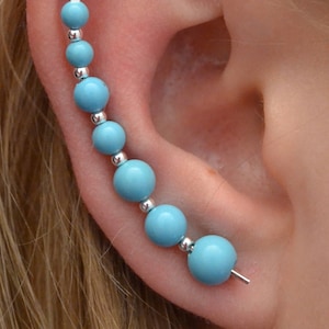 Könnte beinhalten: Ein türkisfarbener Ohrklemme mit einer Reihe von abgestuften, runden Perlen, die durch kleine silberfarbene Perlen verbunden sind. Der Ohrschmuck wird am Ohr getragen, mit einem kleinen Stift zur sicheren Befestigung. Der Schmuck ist vor dem Hintergrund von blondem Haar und Haut platziert.