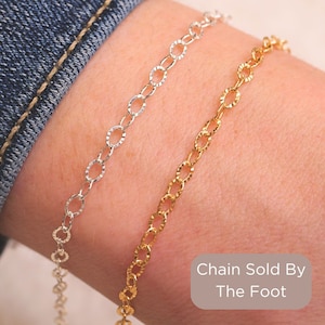 May include: Two chain bracelets, one silver and one gold, on a person's wrist. The bracelets are made of small, oval-shaped links. The text "Chain Sold By The Foot" is visible below the bracelets.