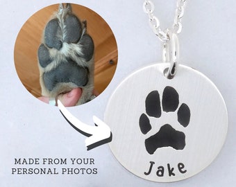 May include: A silver pendant necklace featuring a paw print and the name "Jake". The necklace is connected to a chain. An inset image shows a dog's paw. Text reads "MADE FROM YOUR PERSONAL PHOTOS".