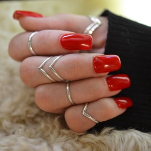May include: A hand wearing multiple silver rings, including a chevron design, with bright red nail polish.