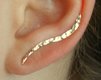 May include: A gold ear cuff with a hammered finish. The cuff is curved and has a pointed end. It is worn on the upper part of the ear.