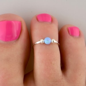 May include: A silver toe ring with a light blue opal-like bead in the center. The ring is worn on a toe with pink nail polish.