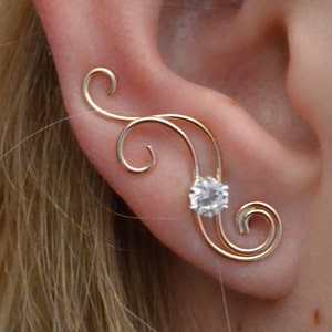 May include: A gold ear cuff with a single clear rhinestone. The ear cuff is shaped like a delicate, swirling vine.