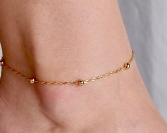 May include: A delicate gold anklet with small round beads. The anklet is on a woman's foot with gold toe rings.