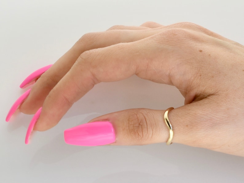 May include: A gold-colored ring with a curved design, worn on the thumb of a hand with pink nail polish.