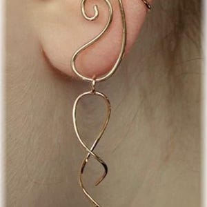 Könnte beinhalten: Ein goldfarbenes Ohr-Manschetten- und Ohrring-Set. Die Ohr-Manschette verläuft über das Ohr, während der Ohrring ein Wirbel- und ein verdrehtes, längliches Design aufweist. Der Schmuck besteht aus dünnem Metalldraht.