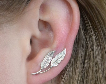 May include: Silver feather ear cuff earrings. The earrings are designed to look like two feathers with a stem connecting them. The earrings are worn on the upper part of the ear.