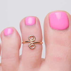 May include: A gold toe ring with a double loop design. The ring is worn on a toe with pink nail polish.