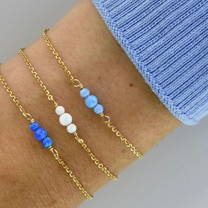Könnte beinhalten: Drei goldene Kettenarmbänder mit unterschiedlich farbigen Opalperlen. Das obere Armband hat zwei hellblaue Opalperlen, das mittlere Armband hat zwei weiße Opalperlen und das untere Armband hat drei blaue Opalperlen.
