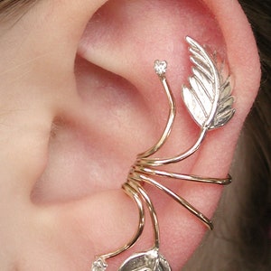 May include: Gold ear cuff with a leaf design and two small clear stones. The cuff wraps around the ear and has a delicate, feminine look.