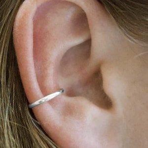 May include: A close-up of a person's ear with a silver ear cuff. The ear cuff is a simple, thin band that wraps around the upper part of the ear.
