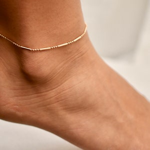 Peut inclure: Un bracelet de cheville doré délicat, composé d'une série de petites perles rondes et de segments en forme de barres allongées. Le bracelet de cheville est porté sur un pied, mettant en valeur son design minimaliste et son élégance subtile. Un accessoire élégant.