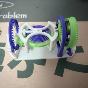 May include: A 3D-printed mechanical model featuring a white base and frame, with purple, green, and blue gears and a central spring. The gears are interlocked, showcasing a complex design. The model is designed for educational or display purposes.