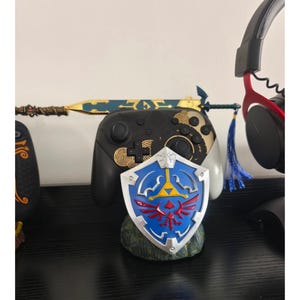 Puede incluir: Se muestra un escudo azul y rojo con un emblema dorado, un mando de juego negro y una espada dorada y azul. También se ve un auricular negro con detalles en rojo. Los objetos están dispuestos sobre una superficie negra.