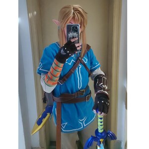 K&ouml;nnte beinhalten: Ein Cosplay-Kost&uuml;m bestehend aus einer leuchtend blauen Tunika mit wei&szlig;em Saum, braunen lederartigen Riemen und G&uuml;rteln. Das Outfit umfasst dunkle fingerlose Handschuhe mit silbernen Akzenten, einen gemusterten Armschutz und ein blau-goldenes Schwert.