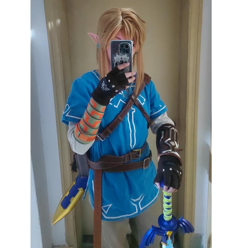 K&ouml;nnte beinhalten: Ein Cosplay-Kost&uuml;m bestehend aus einer leuchtend blauen Tunika mit wei&szlig;em Saum, braunen lederartigen Riemen und G&uuml;rteln. Das Outfit umfasst dunkle fingerlose Handschuhe mit silbernen Akzenten, einen gemusterten Armschutz und ein blau-goldenes Schwert.