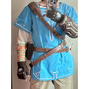 K&ouml;nnte beinhalten: Eine leuchtend blaue Tunika mit wei&szlig;en Stickereidetails, getragen mit einem braunen, lederartigen Schulterriemen und H&uuml;ftg&uuml;rtel. Das Outfit umfasst einen dunkelbraunen fingerlosen Handschuh mit wei&szlig;em Emblem, einen gemusterten Armschutz und eine Armschiene.