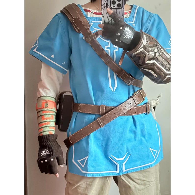 K&ouml;nnte beinhalten: Eine leuchtend blaue Tunika mit wei&szlig;en Stickereidetails, getragen mit einem braunen, lederartigen Schulterriemen und H&uuml;ftg&uuml;rtel. Das Outfit umfasst einen dunkelbraunen fingerlosen Handschuh mit wei&szlig;em Emblem, einen gemusterten Armschutz und eine Armschiene.