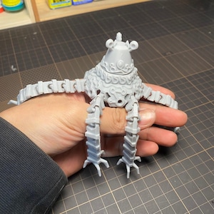 Op de afbeelding: Een grijs, 3D-geprint model van een mechanisch wezen met een afgeronde kop, gesegmenteerde armen en benen. Het model heeft ingewikkelde details en wordt in de hand van een persoon gehouden. Het ontwerp van het wezen doet denken aan een videogame-personage.