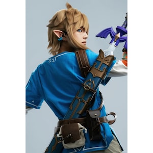 K&ouml;nnte beinhalten: Ein Cosplay-Kost&uuml;m bestehend aus einer leuchtend blauen Tunika mit wei&szlig;em Saum, einem braunen Lederg&uuml;rtel mit angebrachten Taschen sowie einer blau-goldenen Schwertscheide mit einem Schwert, das einen violetten Griff aufweist. Die Figur hat blondes Haar und spitze Ohren.