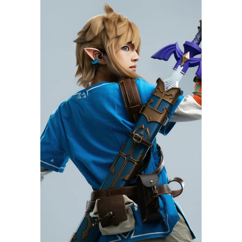 K&ouml;nnte beinhalten: Ein Cosplay-Kost&uuml;m bestehend aus einer leuchtend blauen Tunika mit wei&szlig;em Saum, einem braunen Lederg&uuml;rtel mit angebrachten Taschen sowie einer blau-goldenen Schwertscheide mit einem Schwert, das einen violetten Griff aufweist. Die Figur hat blondes Haar und spitze Ohren.