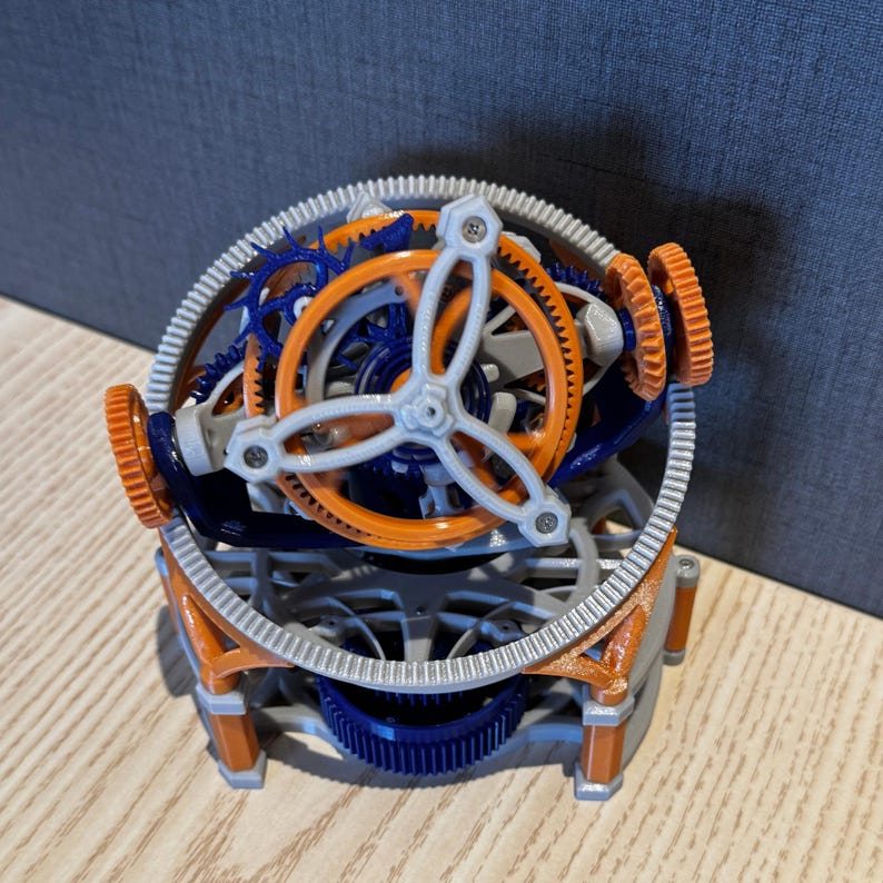 以下が含まれることがあります： オレンジ、青、グレーの歯車が複雑に配置された3Dプリントの機械モデル。中央に白いコンポーネントがあり、オレンジとグレーのベースで支えられた球形デザインです。