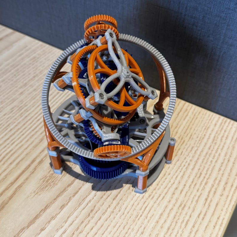 以下が含まれることがあります： オレンジ、青、グレーの複雑な歯車配置が特徴の3Dプリントされたキネティック彫刻。 複雑なデザインには、大きな外側のリングと相互接続された複数の歯車が含まれており、機械的なテーマを示しています。