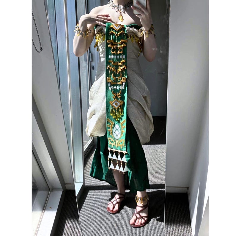 Op de afbeelding: Een strapless cr&egrave;me-kleurige jurk met een donkergroene rok, voorzien van een lange, verticale groene sjerp versierd met ingewikkelde goudkleurige geometrische metalen ornamenten, bungelende kristallen en witte driehoekige accenten, gecombineerd met bijpassende gouden sieraden.