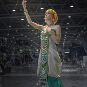 Op de afbeelding: Een kostuum bestaande uit een cr&egrave;mekleurige jurk met een lang, blauwgroen middenpaneel versierd met gouden geometrische patronen en bungelende metalen accenten. De outfit bevat bijpassende gouden armmanchetten, een ketting en puntige oorprotheses.