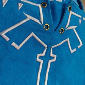 K&ouml;nnte beinhalten: Ein leuchtend blaues Kleidungsst&uuml;ck aus Stoff mit wei&szlig;en, gestickten geometrischen Linienmustern und vier metallisch goldfarbenen &Ouml;sen in V-Ausschnitt-Anordnung. Die Stickerei besteht aus kantigen, symmetrischen Formen am Ausschnitt.