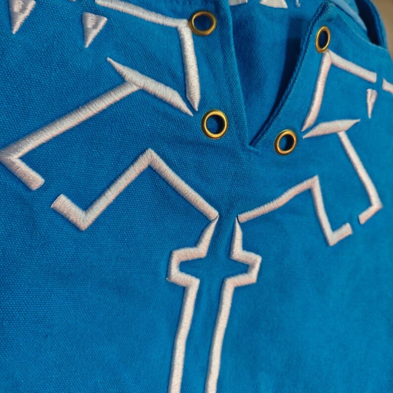 K&ouml;nnte beinhalten: Ein leuchtend blaues Kleidungsst&uuml;ck aus Stoff mit wei&szlig;en, gestickten geometrischen Linienmustern und vier metallisch goldfarbenen &Ouml;sen in V-Ausschnitt-Anordnung. Die Stickerei besteht aus kantigen, symmetrischen Formen am Ausschnitt.