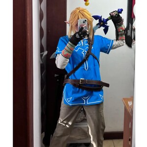 K&ouml;nnte beinhalten: Ein Cosplay-Kost&uuml;m bestehend aus einer leuchtend blauen Tunika mit wei&szlig;em Saum, einer beigen Hose und einem braunen Lederg&uuml;rtel. Das Outfit umfasst schwarze fingerlose Handschuhe mit wei&szlig;en Emblemen, Armschienen und ein blau-silbernes Spielzeugschwert.