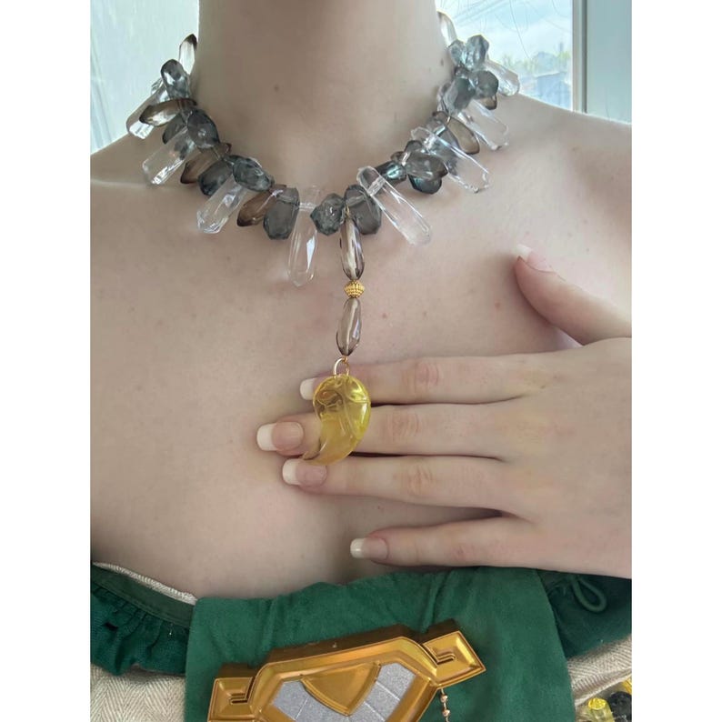 Op de afbeelding: Een ketting met een reeks heldere en rookgrijze kristallen punten in een cirkelvormig patroon. Een goudkleurige kraal verbindt het geheel met een gele, halvemaanvormige hanger met een uitgesneden ontwerp, rustend op de borst.