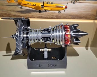 May include: A detailed cross-section model of a jet engine, showcasing internal components in silver and red. The model sits on a black base labeled "TR 900" and "Toy". A yellow airplane painting is in the background.
