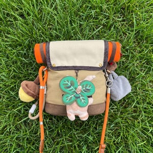 May include: A tan and cream canvas crossbody bag with a fold-over flap, dark brown trim, and bright orange straps. The bag is decorated with a plush acorn charm, a small grey bag charm, and a central green four-leaf clover character charm.