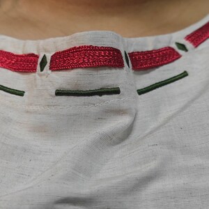 K&ouml;nnte beinhalten: Ein wei&szlig;es Kleidungsst&uuml;ck aus Stoff mit einem runden Halsausschnitt und horizontaler Stickerei. Das Design umfasst ein dickes, strukturiertes rotes Stickmuster, das durch kleine dunkelgr&uuml;ne Rauten und d&uuml;nne dunkelgr&uuml;ne Linien betont wird.