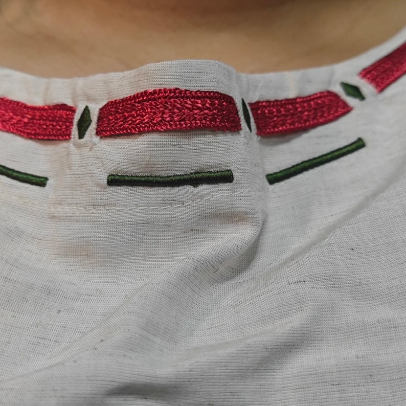 K&ouml;nnte beinhalten: Ein wei&szlig;es Kleidungsst&uuml;ck aus Stoff mit einem runden Halsausschnitt und horizontaler Stickerei. Das Design umfasst ein dickes, strukturiertes rotes Stickmuster, das durch kleine dunkelgr&uuml;ne Rauten und d&uuml;nne dunkelgr&uuml;ne Linien betont wird.
