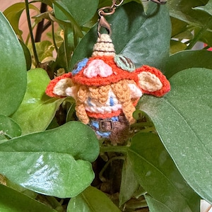 May include: A handmade crochet mushroom charm with a copper-colored chain. The mushroom has a red cap with cream and blue accents, and a small belt. The charm is surrounded by green leaves.