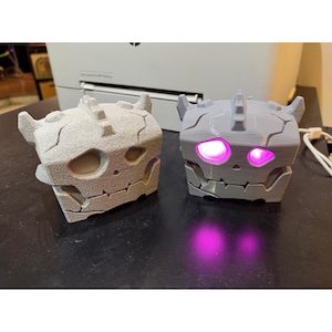 May include: Two square, block-shaped creature figures with horns and jagged, cracked surface details. The left figure is a textured, matte light grey. The right figure is a smooth, light grey plastic with bright, glowing pink LED eyes.