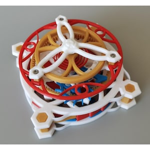 May include: A detailed mechanical model with a layered design. It features red, white, yellow, blue, and gold components, including gears and a central white structure with three arms. The model's design is intricate and colourful, showcasing a complex arrangement.