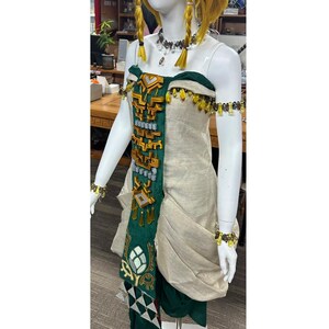 Op de afbeelding: Een beige mouwloze jurk met een centraal donkergroen paneel versierd met gouden en grijze geometrische decoraties. Het kledingstuk heeft een donkergroene halslijn, bijpassende armbanden met gele en grijze kralen en een kralenketting.