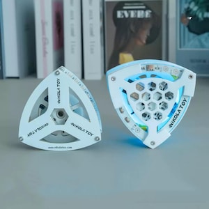 Puede incluir: Dos luces LED triangulares blancas con el texto "NIKOLA TOY" y "www.nikolatoy.com". Una luz está apagada, la otra está iluminada con luz azul. Las luces tienen un patrón de panal y están sobre una superficie gris.