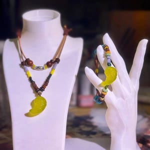 Pode incluir: Um conjunto de colar e pulseira com um pendente amarelo-esverdeado. O colar tem um cordão de couro castanho com contas de madeira e um pendente amarelo-esverdeado. A pulseira tem contas coloridas e um pendente a condizer. Ambos são exibidos em manequins brancos.